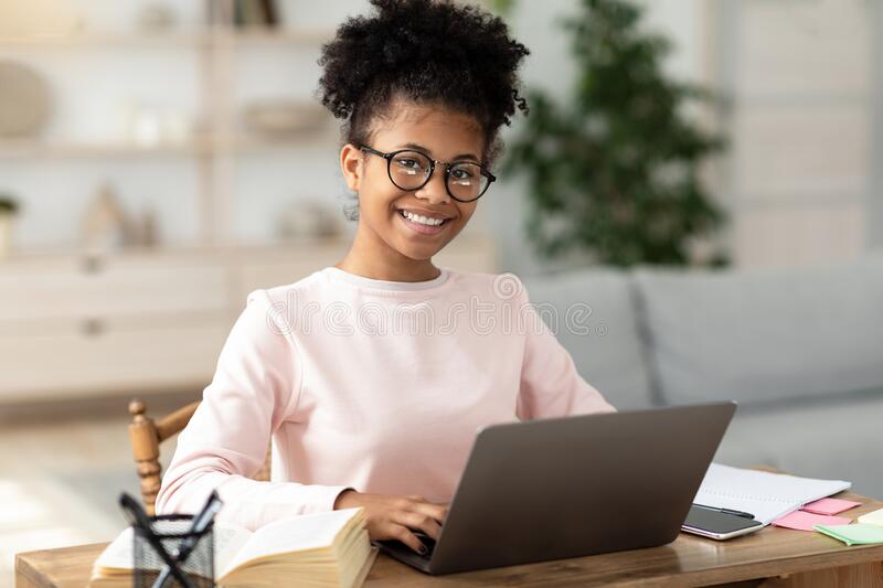black-teen-girl-laptop-learning-online-sitting-home-smiling-to-camera-table-wearing-eyeglasses-distant-school-education-196125818