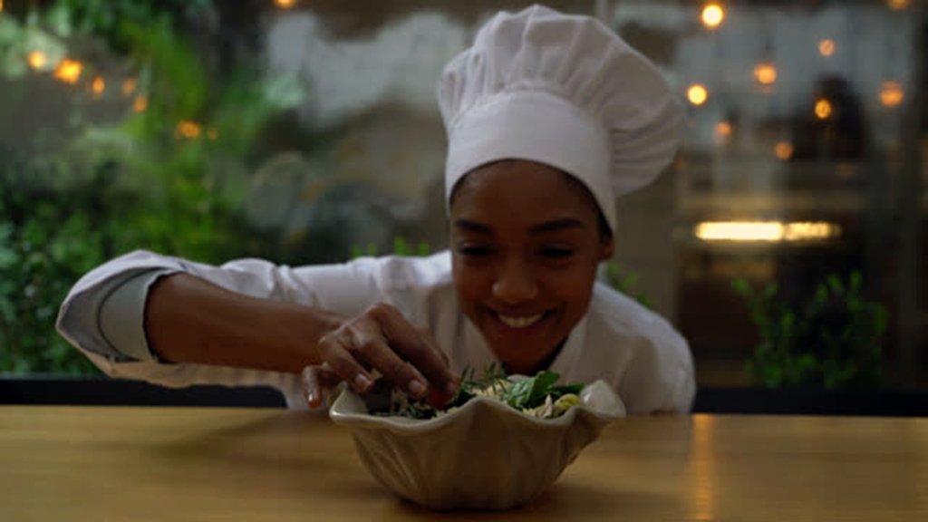 Beautiful black female chef adding a finishing touch to a salad while facing camera smiling very happy
